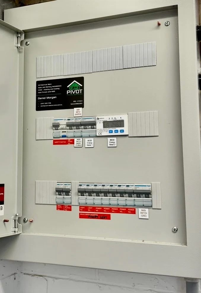 Open switchboard with every circuit labelled, safety switches, two battery energy meters and the Pivot Trade Services contractor plate